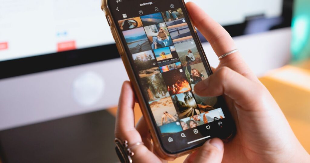 Person browsing an Instagram feed on a smartphone at a desk with a computer monitor behind them.