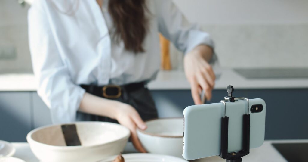Content creator filming a short cooking video on a smartphone mounted on a tripod in a bright kitchen.