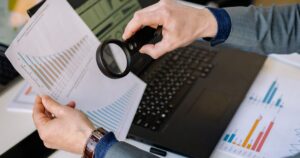 Businessman examining data charts with a magnifying glass over a laptop, representing an SEO audit