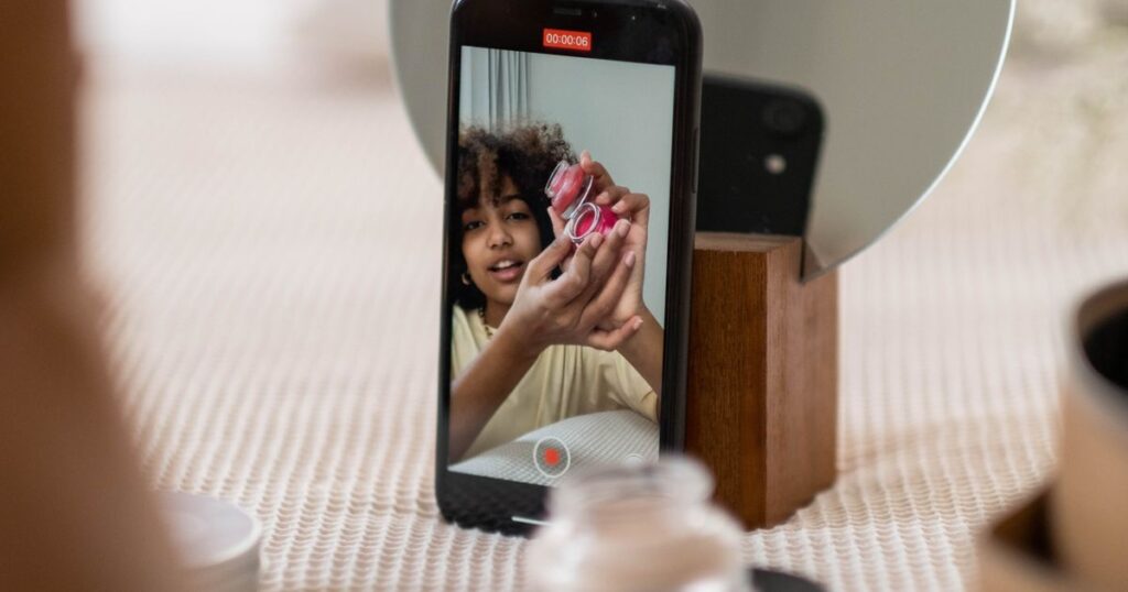 Creator filming a product review on a smartphone while holding a beauty product at a desk.