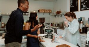 Two returning customers chatting with a barista at a cafe counter, representing repeat custom