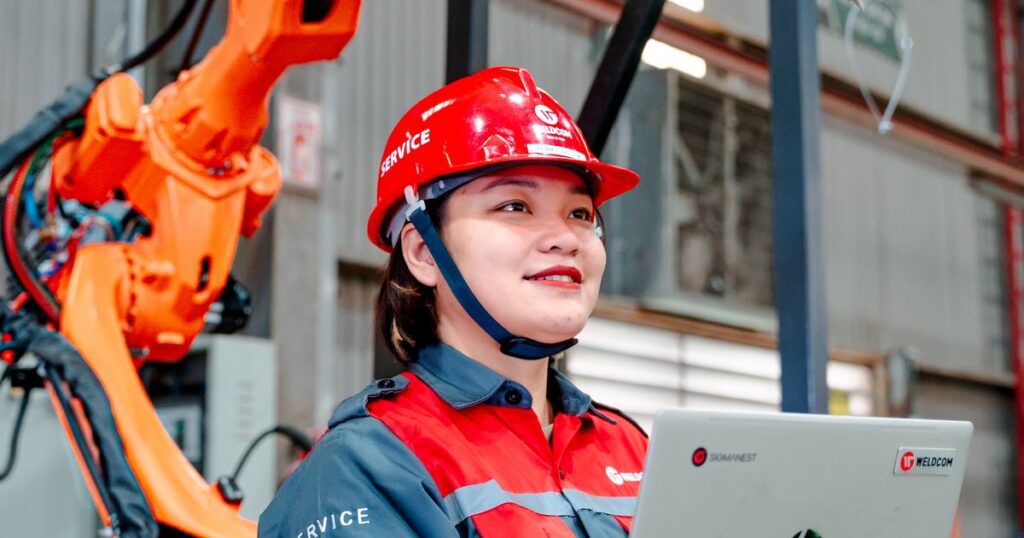 Engineer holding a laptop while supervising a robotic arm on a production line, illustrating human oversight of automated work.