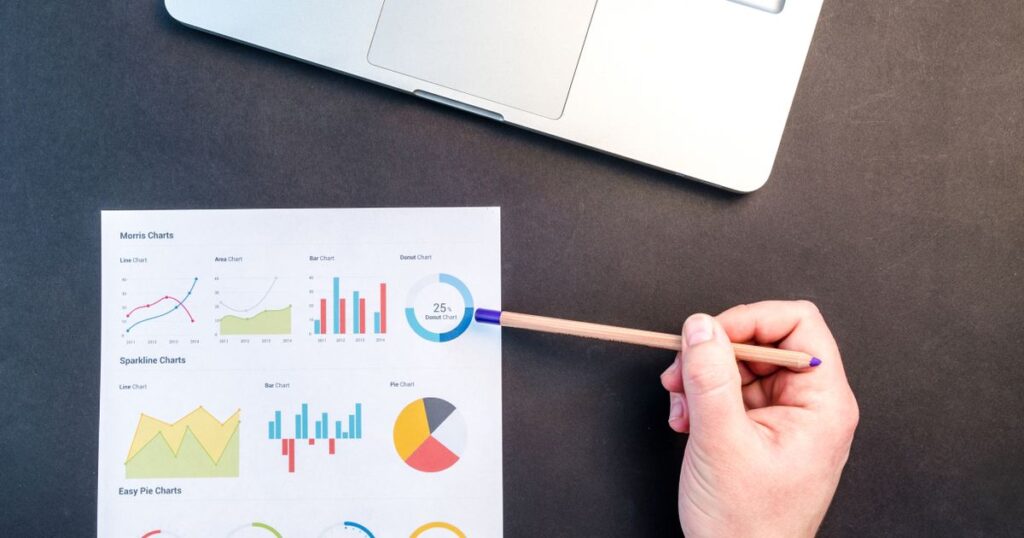 A hand pointing a pencil at a printed sheet of charts on a desk beside a laptop
