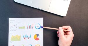 A hand pointing a pencil at a printed sheet of charts on a desk beside a laptop