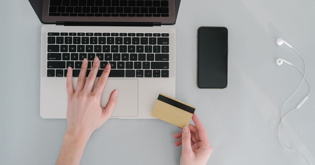 Overhead view of a laptop, a blank payment card, a smartphone and earphones on a pale desk during an online checkout