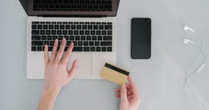 Overhead view of a laptop, a blank payment card, a smartphone and earphones on a pale desk during an online checkout