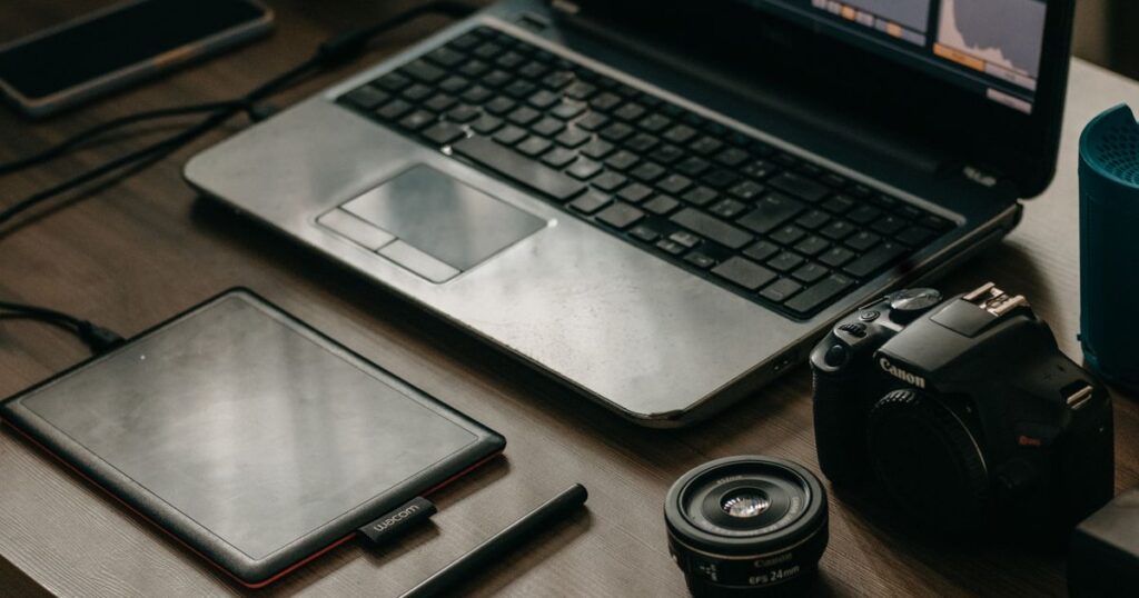 A laptop showing a video editing timeline beside a graphics tablet, stylus, camera body and lens on a desk