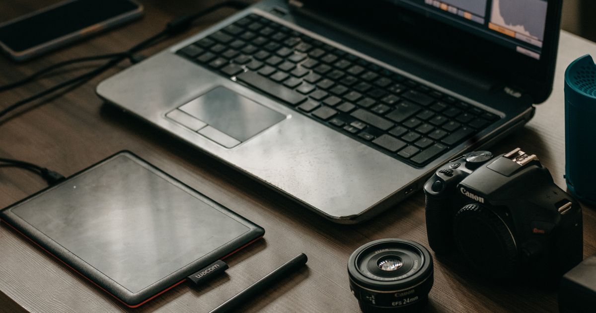 A laptop showing a video editing timeline beside a graphics tablet, stylus, camera body and lens on a desk