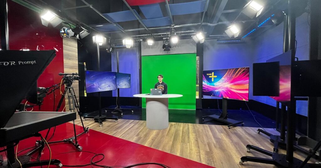 A corporate video studio in use, with a broadcast camera on a tripod, teleprompter, overhead lighting rig and a green screen behind a presenter at a desk.