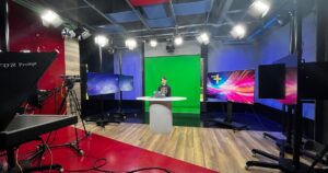 A corporate video studio in use, with a broadcast camera on a tripod, teleprompter, overhead lighting rig and a green screen behind a presenter at a desk.