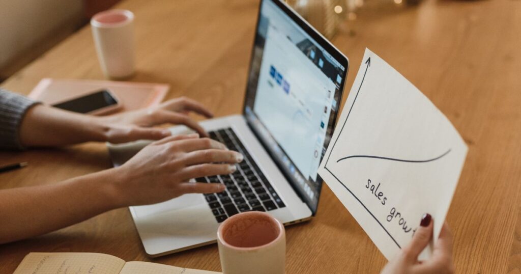 Two people reviewing a hand-drawn sales growth chart beside a laptop at a wooden desk