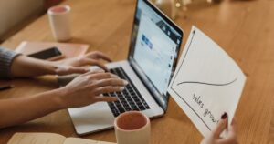 Two people reviewing a hand-drawn sales growth chart beside a laptop at a wooden desk