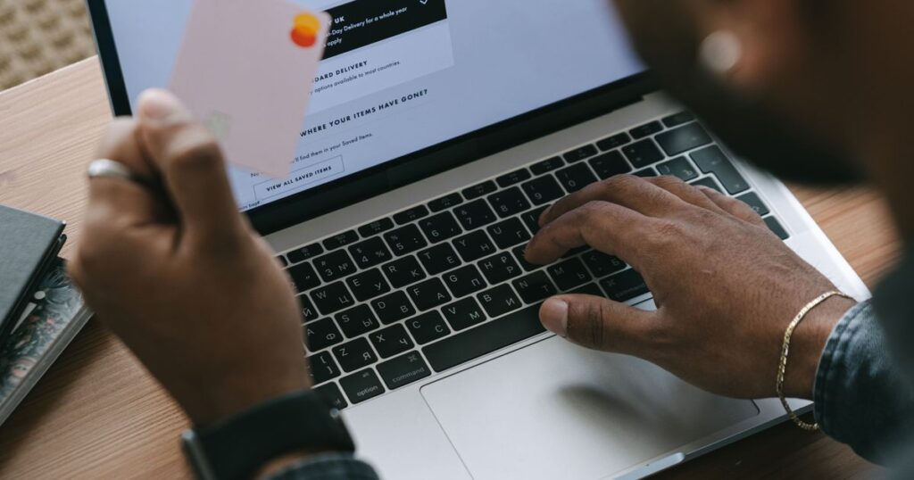 A shopper at a laptop holding a payment card while an online store delivery and cart page is open on screen