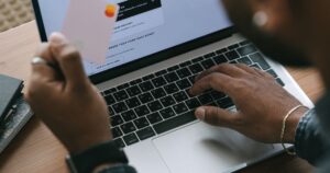 A shopper at a laptop holding a payment card while an online store delivery and cart page is open on screen