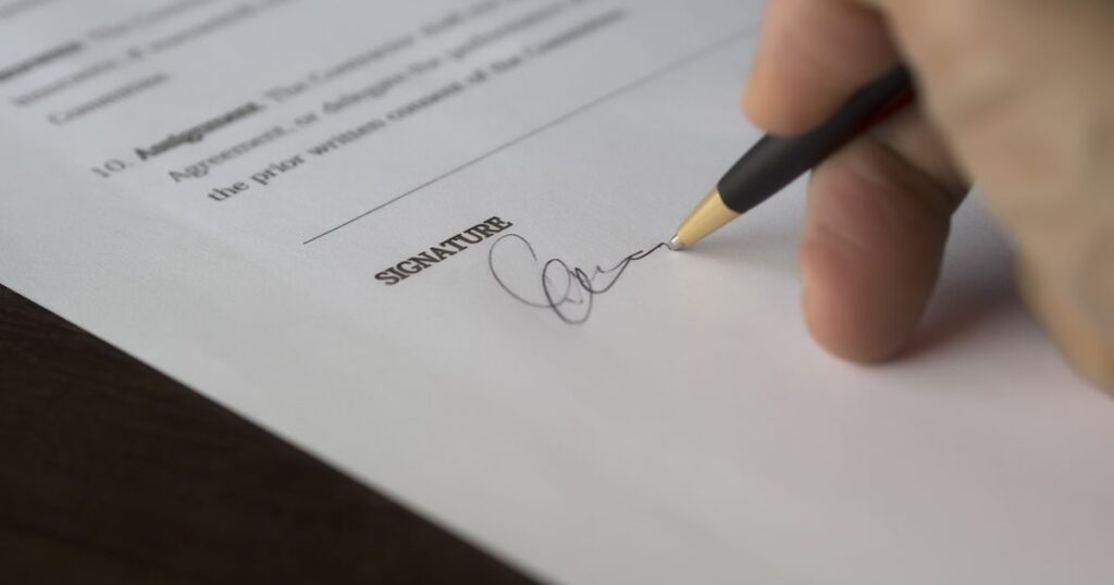 A hand signing a printed document on the signature line with a pen