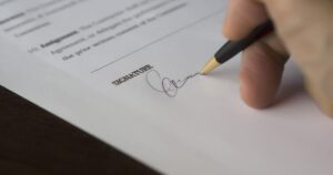A hand signing a printed document on the signature line with a pen