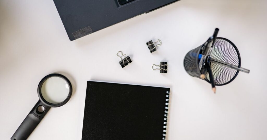 Black notebook, magnifying glass and pens laid out on a white desk beside a laptop while planning keyword research