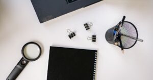 Black notebook, magnifying glass and pens laid out on a white desk beside a laptop while planning keyword research