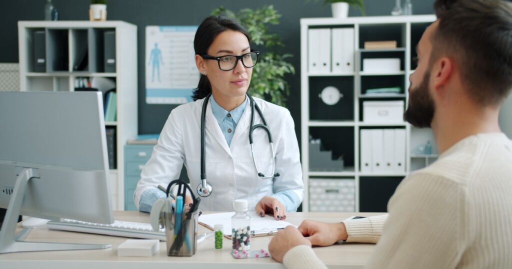 A doctor in a white coat and stethoscope talking with a patient across a desk in a modern clinic consultation room