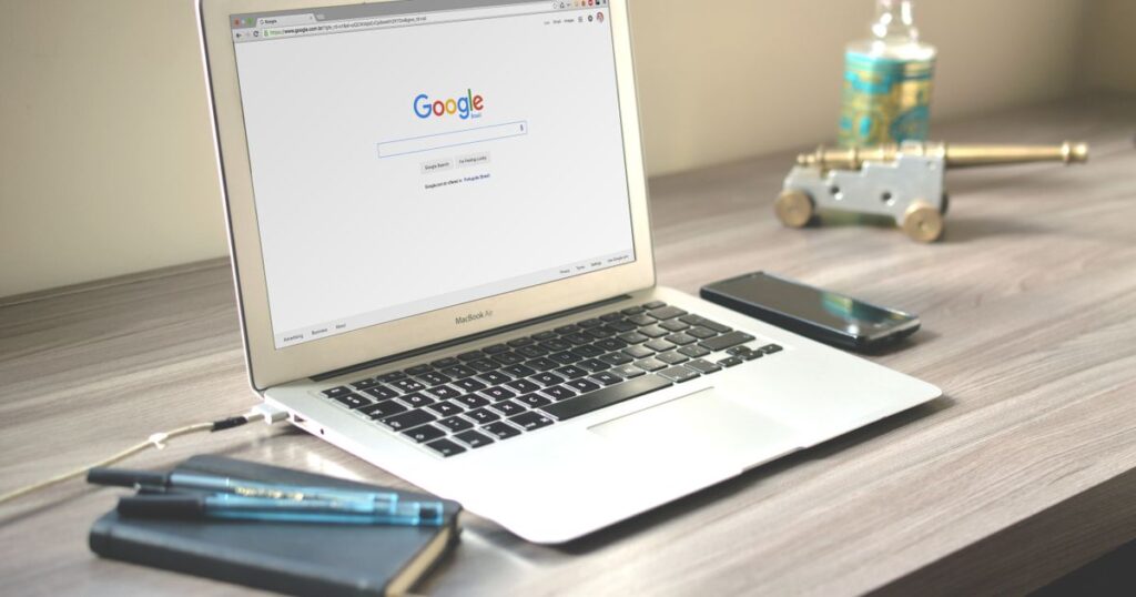 Laptop on a desk displaying the Google search homepage beside a notebook and phone