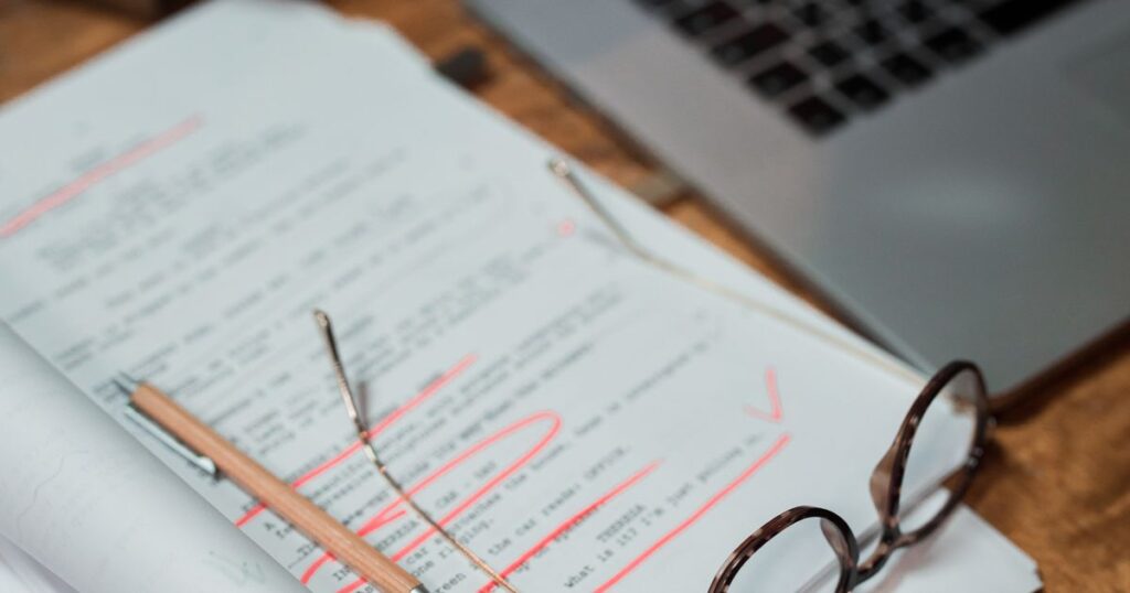 Printed draft marked up in red pen beside reading glasses and a laptop on a wooden desk