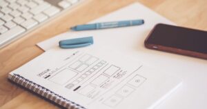 Hand-drawn landing page wireframe sketched in a spiral notebook beside a keyboard, pen and smartphone