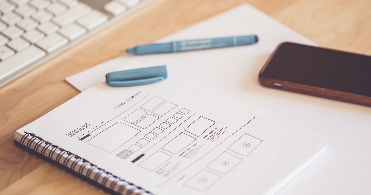 Hand-drawn landing page wireframe sketched in a spiral notebook beside a keyboard, pen and smartphone