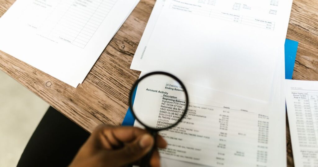 A hand holding a magnifying glass over printed financial statements spread across a desk