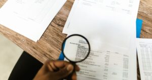 A hand holding a magnifying glass over printed financial statements spread across a desk