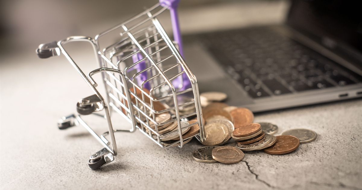 A miniature shopping trolley tipped over beside a laptop, spilling coins across a desk