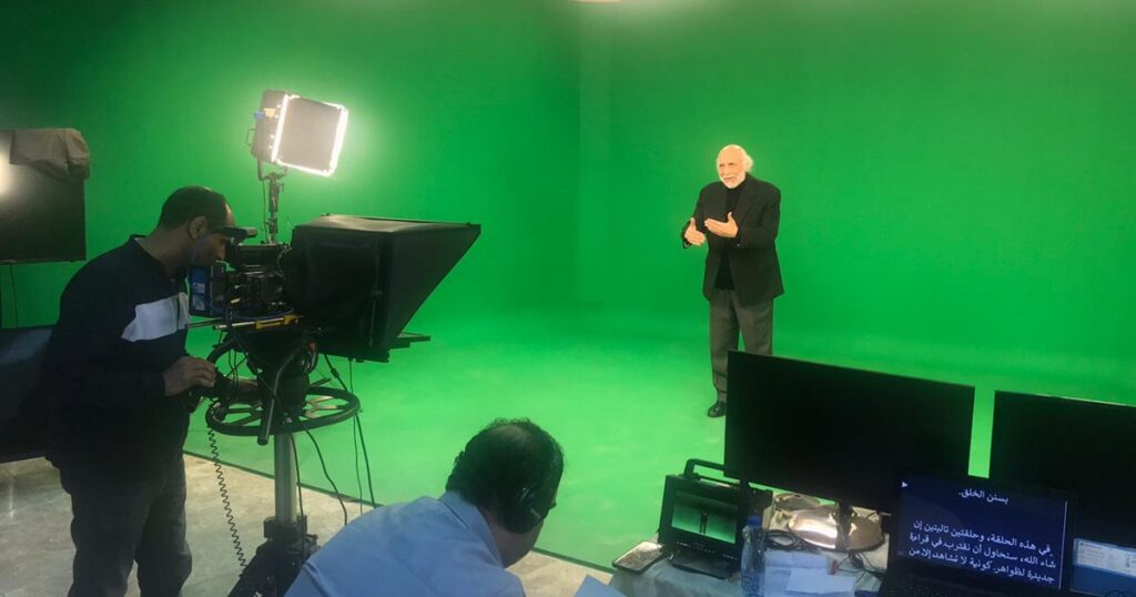 A green-screen studio shoot in progress, with a camera operator, a teleprompter and a director at a monitor while a presenter speaks to camera.