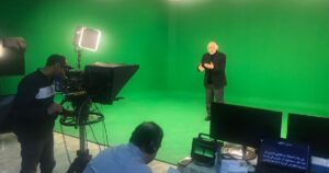 A green-screen studio shoot in progress, with a camera operator, a teleprompter and a director at a monitor while a presenter speaks to camera.