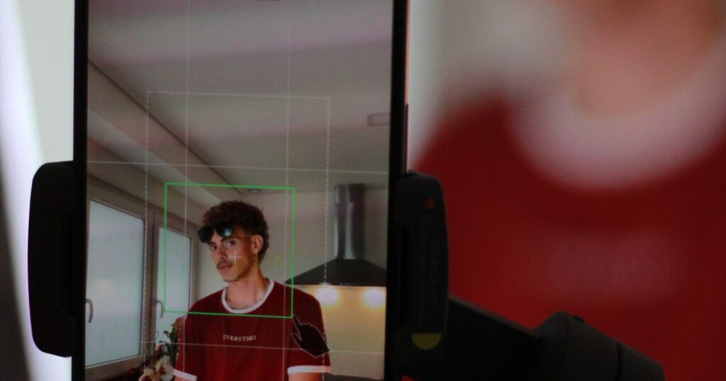 A smartphone mounted on a stand filming a person vertically, with the framing grid and face tracking visible on screen