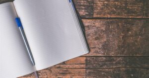 An open lined notebook with a pen resting on it, on a weathered wooden desk, ready for scripting