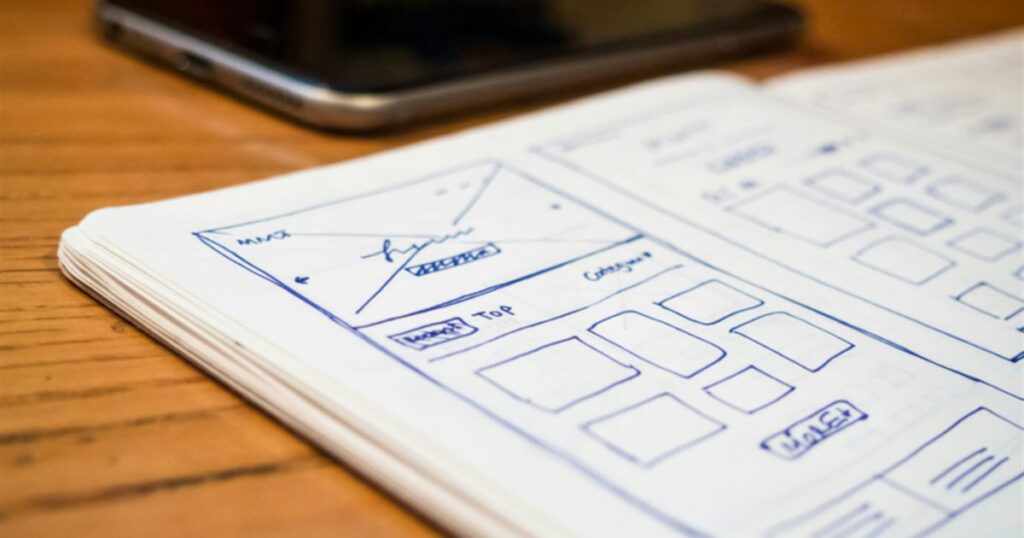 Hand-drawn website wireframe sketches in a notebook beside a smartphone on a wooden desk