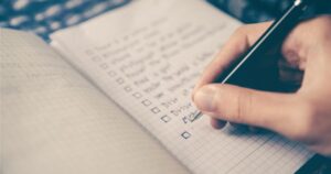 Person ticking off items on a handwritten checklist in a grid notebook with a pen