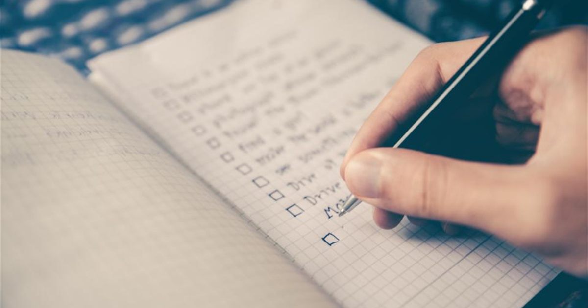 Person ticking off items on a handwritten checklist in a grid notebook with a pen