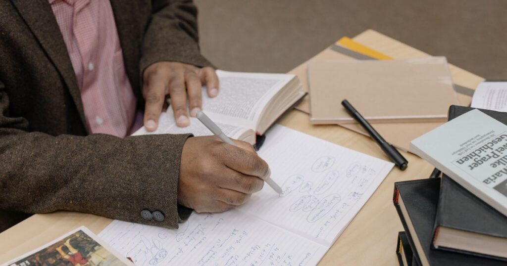 Researcher taking handwritten notes from an open book at a desk stacked with papers