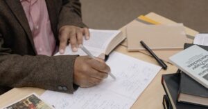 Researcher taking handwritten notes from an open book at a desk stacked with papers