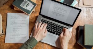 Writer typing a draft on a laptop beside a printed manuscript on a wooden desk