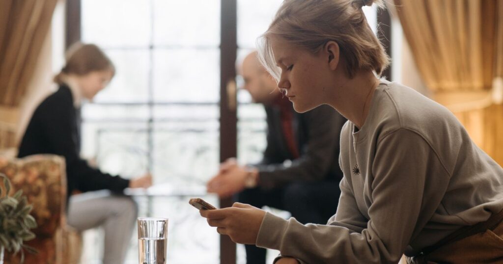 A teenager looking at a smartphone in a living room with adults blurred in the background