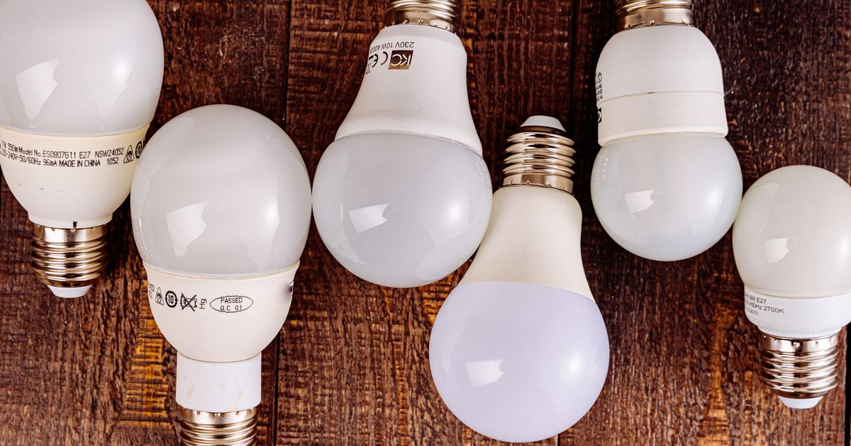 Seven unbranded LED and compact fluorescent light bulbs on a wooden table, showing their printed wattage and compliance markings.