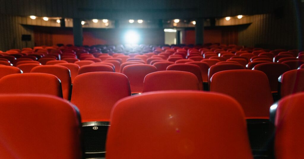 Rows of empty red seats in a dark cinema, facing a bright projector light at the back of the hall.