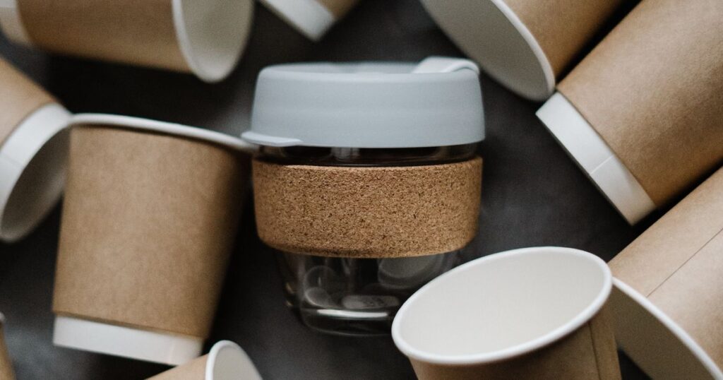 A reusable glass cup with a cork band surrounded by disposable paper cups, illustrating environmental claims in product marketing.