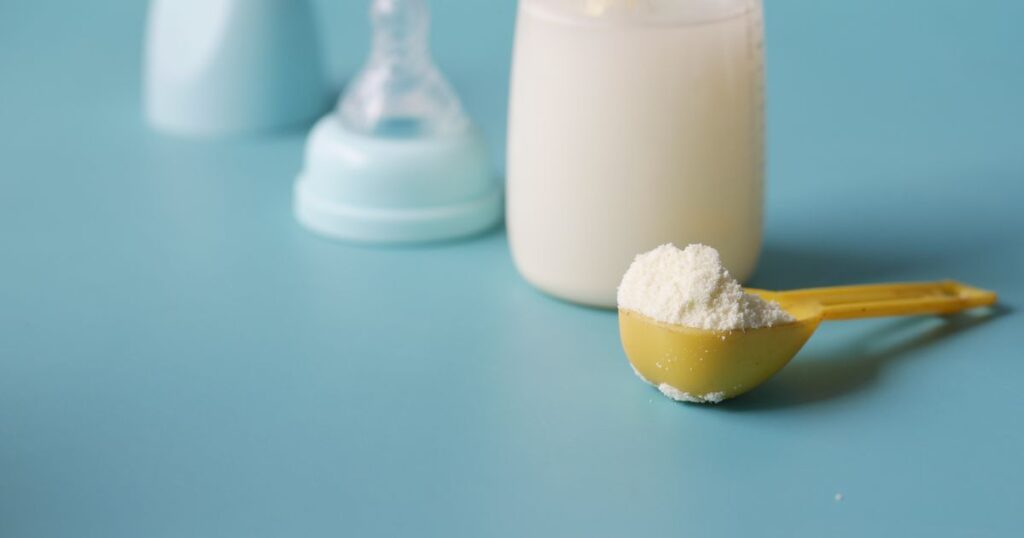 A yellow scoop of milk formula powder beside a feeding bottle and teat on a pale blue surface.