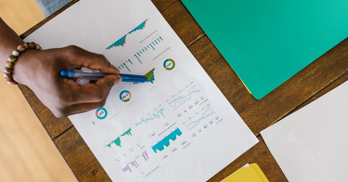 A hand marking printed business charts and financial figures with a pen on a wooden desk, as in preparing investor materials.