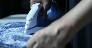 Close-up of hands ironing a blue patterned shirt, representing domestic work in an article on maid agency advertising rules in Singapore.