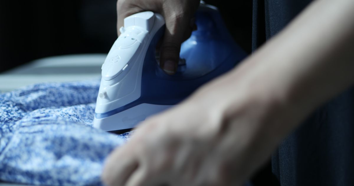 Close-up of hands ironing a blue patterned shirt, representing domestic work in an article on maid agency advertising rules in Singapore.
