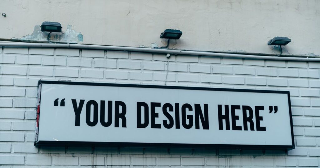 A blank white signboard mounted on a painted brick wall under three floodlights, the kind of outdoor sign that needs a licence in Singapore.