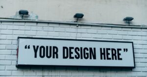 A blank white signboard mounted on a painted brick wall under three floodlights, the kind of outdoor sign that needs a licence in Singapore.